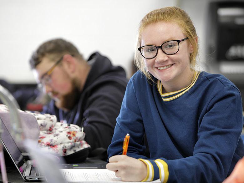 female student with red hair and glasses in a classroom holding a pen and looking at the camera; male student in the background looking down
