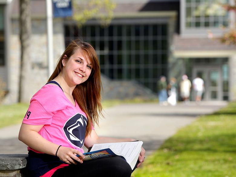 Young woman in a bright Penn State t-shirt sitting on the wall in front of Hayfield House studying