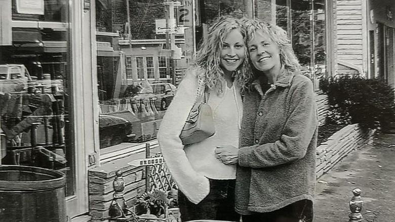 A black and white photo of two woman standing outside a business with their arms around each other.