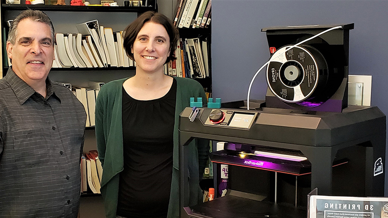 Jonathan Pineno (Friedman Art Gallery director) and Megan Mac Gregor (librarian) standing in front of the Nesbitt Library's 3D printer; two of the small green widgets are sitting on top.
